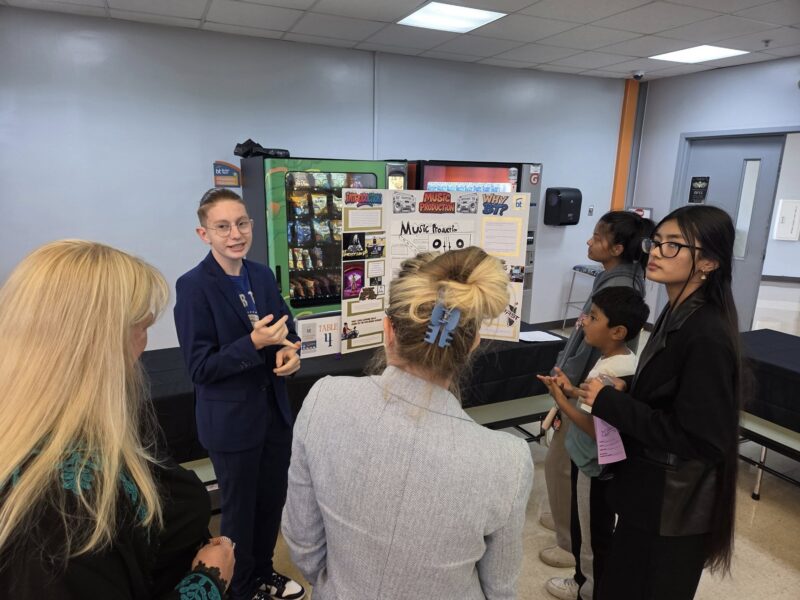 A 10th grade student in a blue suit presents a science project display to a group of people, including adults and children, in a classroom or academy setting. The display board behind the student contains colorful images and text for career exploration.