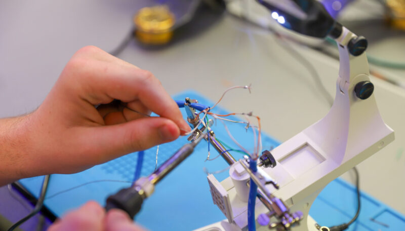 A person uses a soldering iron and a helping hand tool to connect wires on a workbench, focusing on electronics assembly or repair tasks vital to Information Technology projects.