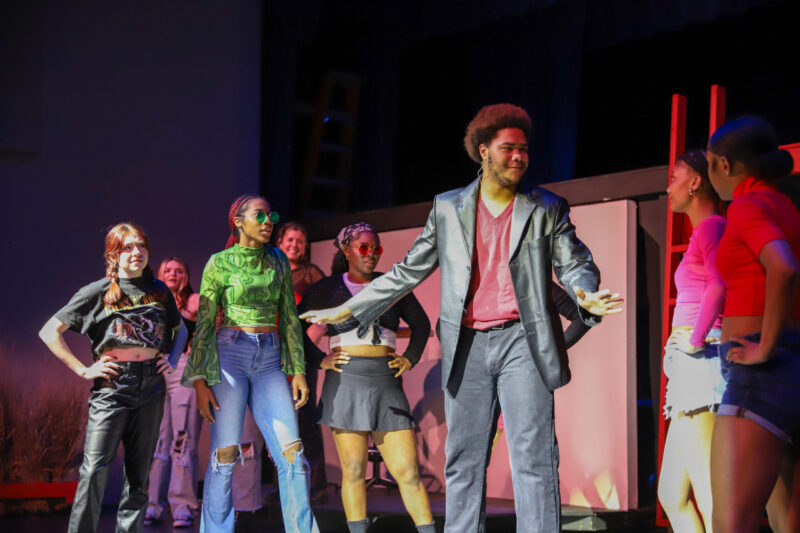 A group of young performers stands on stage under bright lights, with one person in a dark blazer gesturing confidently while others watch and smile, suggesting a lively theatrical scene.