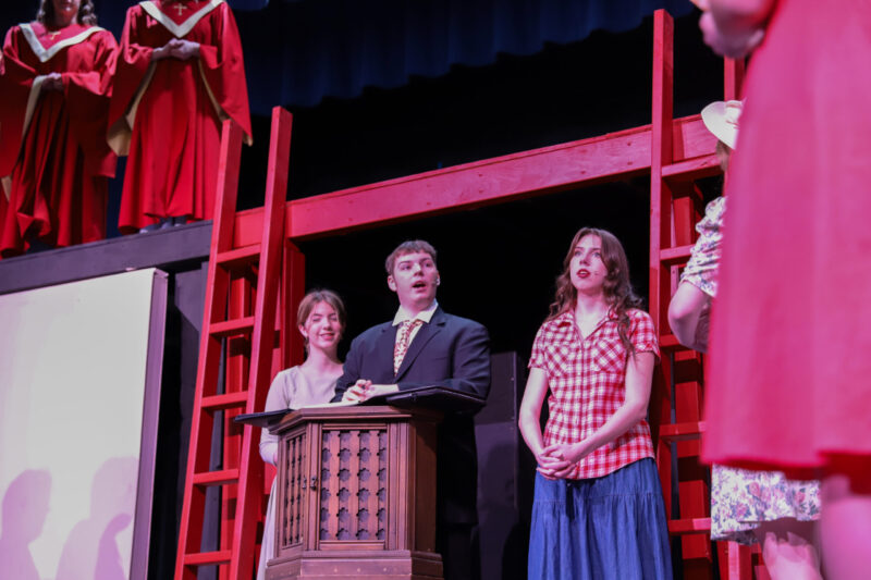 A group of people perform on stage; a man in a suit stands at a podium, flanked by two women, with others in red robes above and more performers in colorful clothes nearby. The set features bright red ladders and beams.