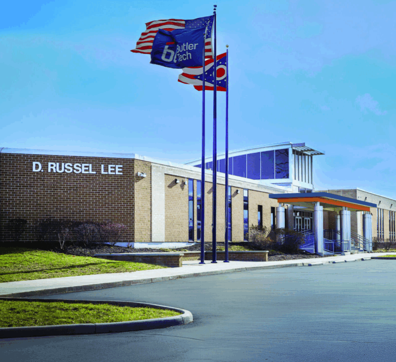 The image shows the D. Russel Lee building at Butler Tech, with its modern design and large windows. Three flagpoles display the American, Butler Tech, and Ohio flags outside on a clear day near the paved entrance area.