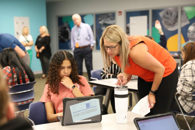 A young student sits at a desk, focusing on a laptop. An adult woman stands next to her, engaging and pointing at the screen, providing assistance. Several other people are visible in the background, interacting and standing around in a classroom setting as Butler Tech launches its new Business Program.