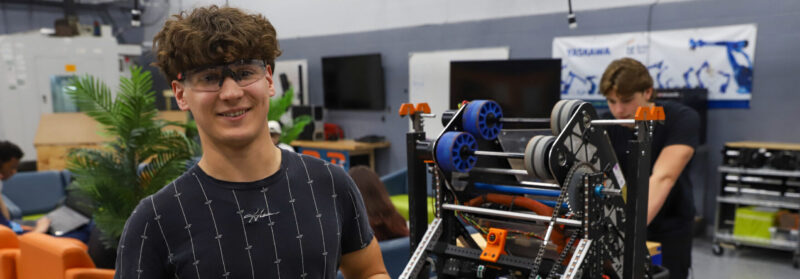 A smiling engineering student wearing safety glasses holds metal parts in a robotics lab, standing next to a large, partially assembled robot. Background shows other students in this dynamic 3-year program working amid equipment on tables.