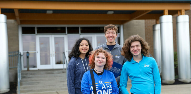 Four young people stand together outside a building. They are smiling and dressed in casual attire, with two of them wearing blue shirts featuring a logo and text promoting Senior Programs. The building entrance behind them has glass doors and a covered walkway.