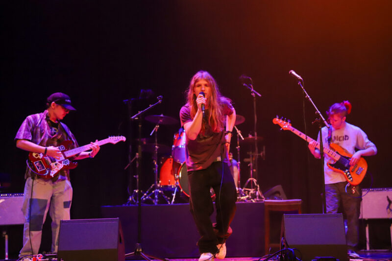 A band performs on stage under colorful lights; the lead singer sings into a microphone while two guitarists play on either side, with drums in the background—capturing the dynamic energy of live music and Recording Arts.
