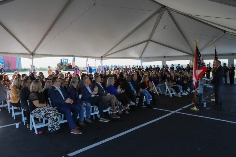 A crowd of people seated under a large white tent listens attentively to a speaker at a lectern adorned with the United States and state flags. The audience, including men and women in business attire, gathers to mark the expansion of Butler Tech's Bioscience Center. A construction vehicle is visible in the background.