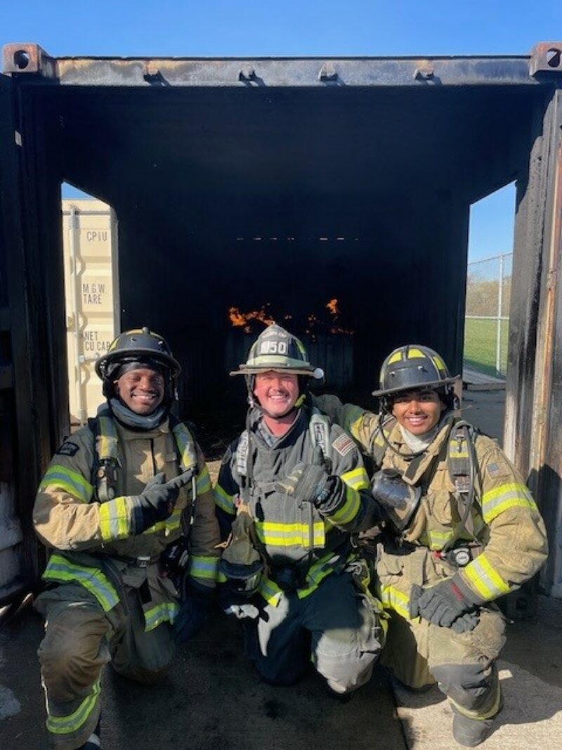 Three firefighters, all Butler Tech Alumni in full gear, including helmets and protective suits, smile while crouching in front of a container with a small, controlled fire inside. The outdoor setting is bright and clear.
