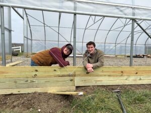 Two students lean on the wooden frame of a greenhouse under construction. Both are wearing jackets and smiling at the camera. The structure, a testament to green engineering, features metal frames and clear panels. Tools are scattered around, transforming waste into wealth with every bolt and beam.