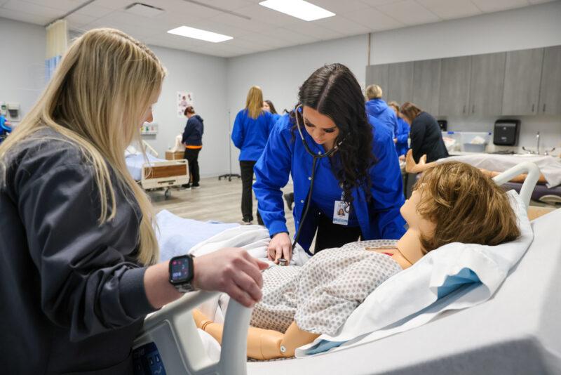 In the Butler Tech lab, nursing students in the RN program practice clinical skills on a medical mannequin. Kayla Bard uses her stethoscope on its chest, with one peer observing and others working in the background, all clad in blue scrubs.