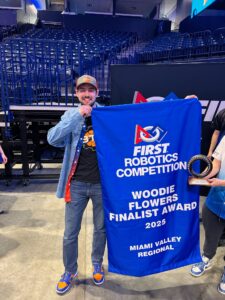 A person smiling and holding a blue banner that reads "FIRST Robotics Competition, Woodie Flowers Finalist Award 2025, Miami Valley Regional." They are also holding a circular award. The background features stadium seating.
