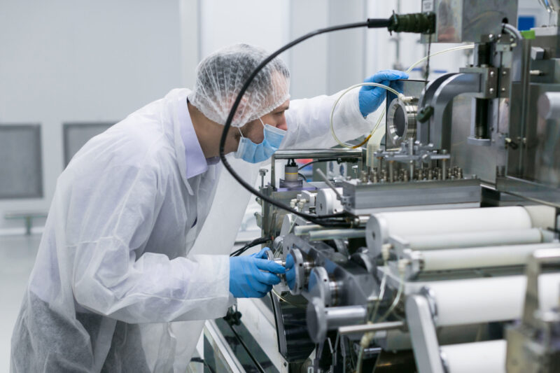 A person in protective clothing, a hairnet, mask, and gloves operates or inspects complex machinery in a clean, sterile laboratory or GMP-compliant manufacturing environment.