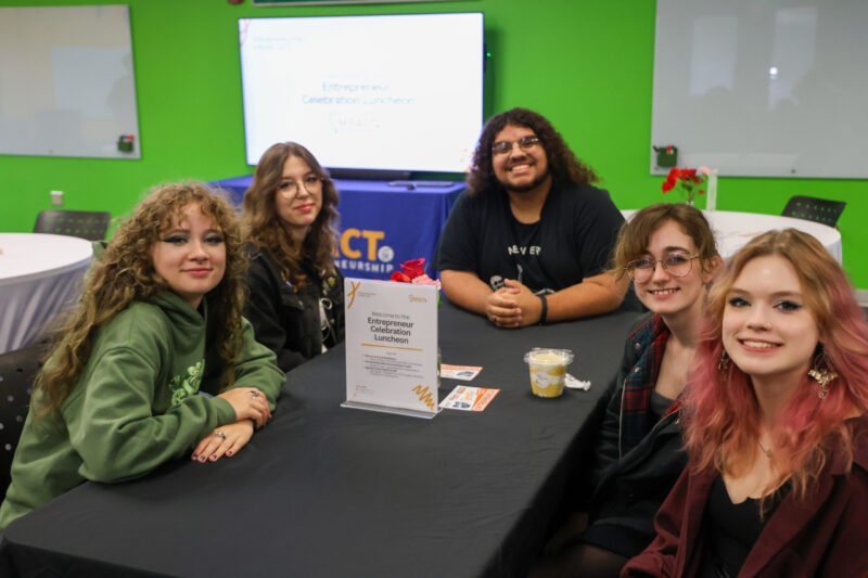 Five young student entrepreneurs sit around a table with a black tablecloth at the Entrepreneur Celebration Luncheon. Hosted by the Greater Hamilton Chamber of Commerce, all are smiling and facing the camera, ready to receive their gift.