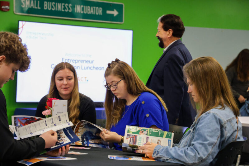 Four people sit at a table looking through magazines or brochures, while another stands nearby. A sign reads