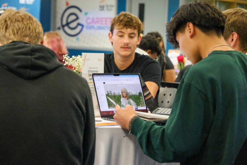 A student in a green sweater works on a laptop displaying an image of a woman in a field, while other student entrepreneurs gather around a table in a classroom with a