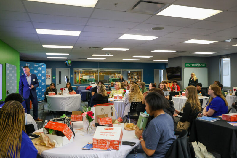 A group of people sit at round tables with pizza boxes in a brightly lit room, listening to a man speaking at the front near a blue Greater Hamilton Chamber of Commerce banner. The setting appears to be a community or networking event for student entrepreneurs.