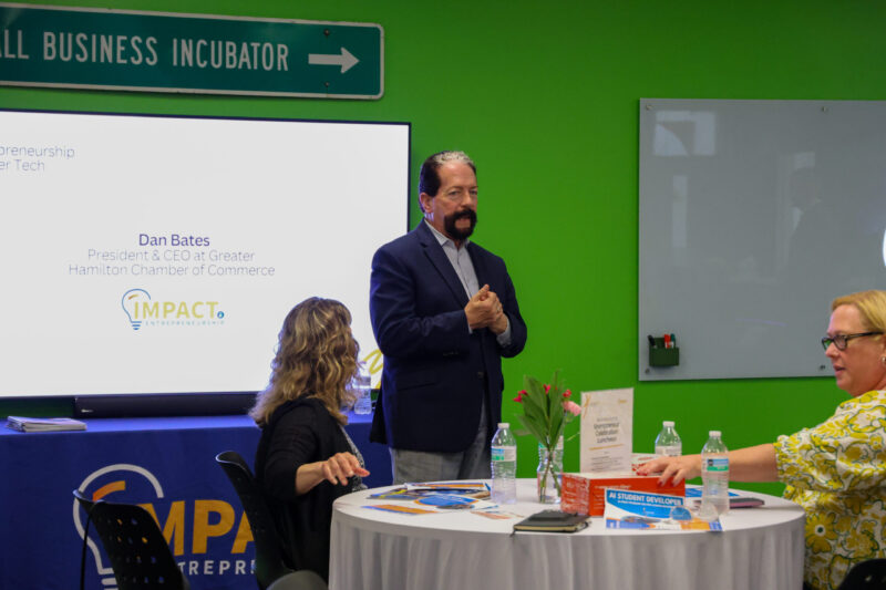 A man in a suit speaks before a screen displaying his name and title at a business event for the Greater Hamilton Chamber of Commerce, while two seated women listen attentively at a round table set with water bottles.