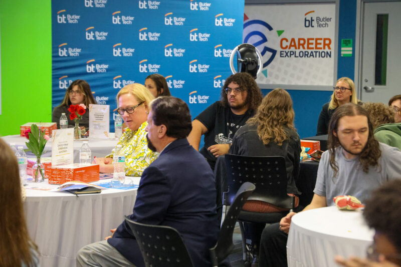 A group of people, including student entrepreneurs, sit at round tables in a room decorated with Butler Tech banners. Some are eating and talking, while a sign in the background reads “Career Exploration.”.