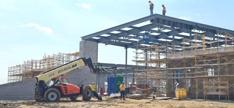 Construction site at Butler Tech Aviation Center: workers and scaffolding surround steel beams as a forklift operates below; two people stand on the roof, marking the Topping Out Milestone under a clear sky.