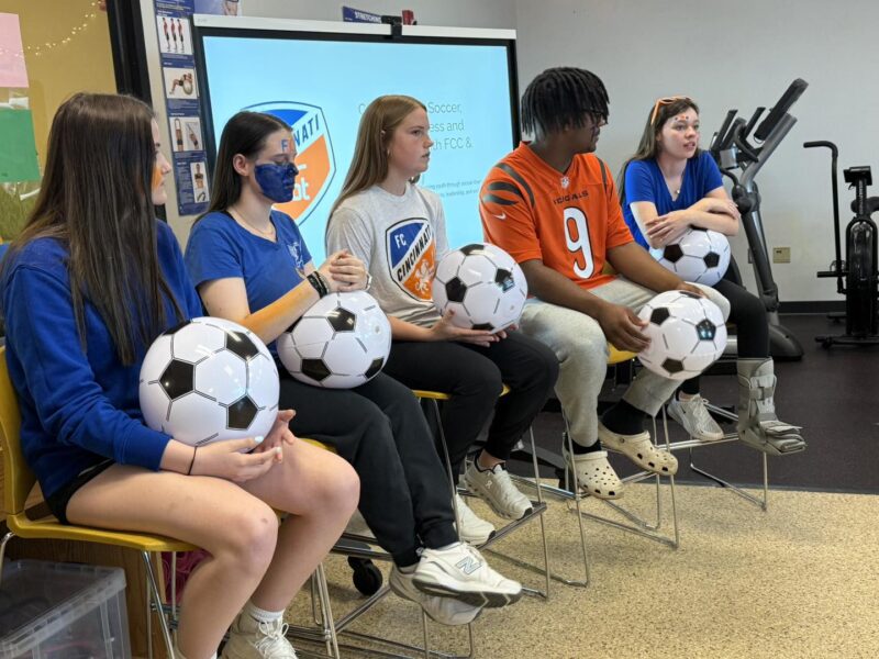Five young people sit in a row holding soccer balls, engaged in conversation. Four wear blue sportswear, one wears an orange striped jersey. Exercise equipment and a screen are visible in the background.
