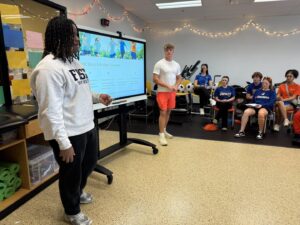 Two students stand and present in front of a large screen displaying a slideshow to a group of seated classmates in a classroom decorated with string lights. Some students wear sports attire and hold notepads.