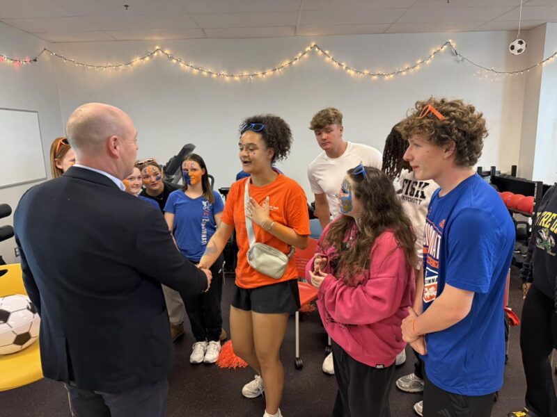 A man in a suit shakes hands with a smiling young woman in a group of casually dressed students, some wearing sports shirts and face paint, in a decorated room with fairy lights and sports equipment.