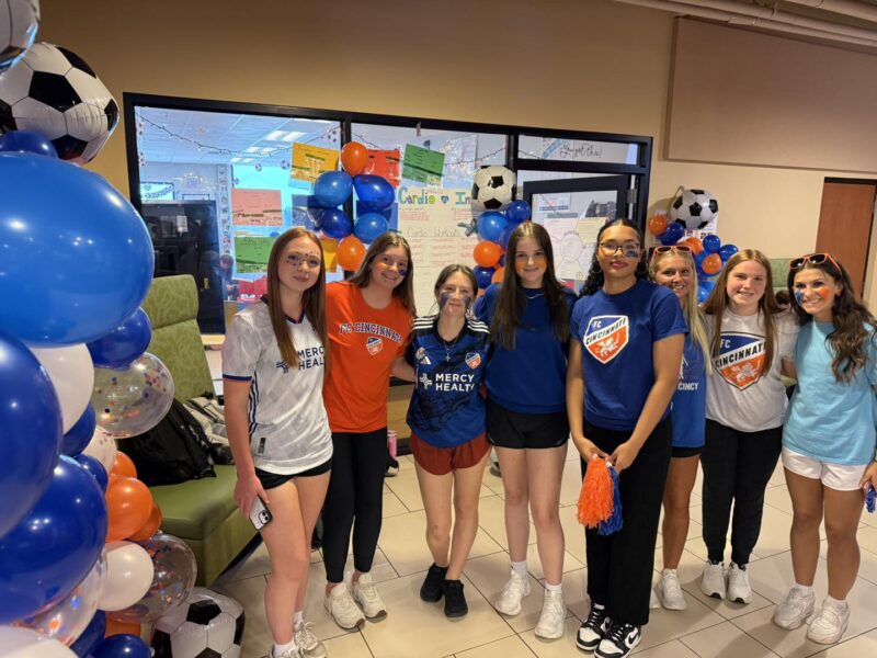 A group of eight smiling girls stands indoors, surrounded by blue, white, and orange soccer-themed balloons and decorations. They are wearing soccer shirts and shorts, posing for a team photo at an event.