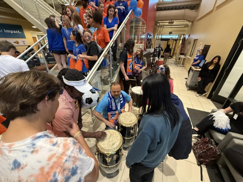 A group of people gather indoors near a staircase. Some are playing drums while others, wearing colorful shirts and scarves, stand on the stairs holding balloons and cheering, creating a lively, festive atmosphere.