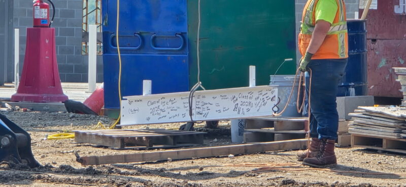 A construction worker stands near a signed steel beam at an Aviation Center worksite, celebrating the Topping Out Milestone. Wearing a safety vest, gloves, and boots, he surveys the Butler Tech project as equipment and materials lie scattered nearby.