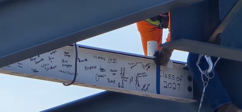 A construction worker in an orange shirt secures a steel beam covered in handwritten signatures and messages, marking the Topping Out Milestone for Butler Tech's new Aviation Center and honoring the