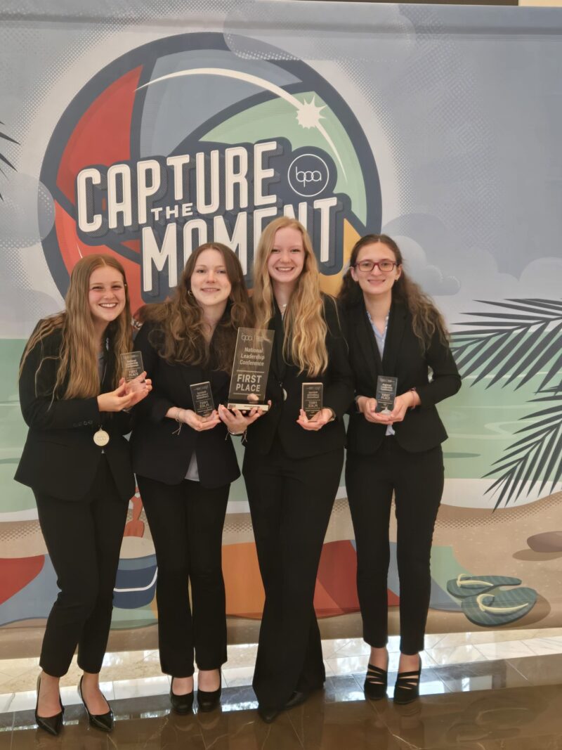 Four young women in business attire stand smiling and holding trophies as BPA National Champions in front of a colorful 