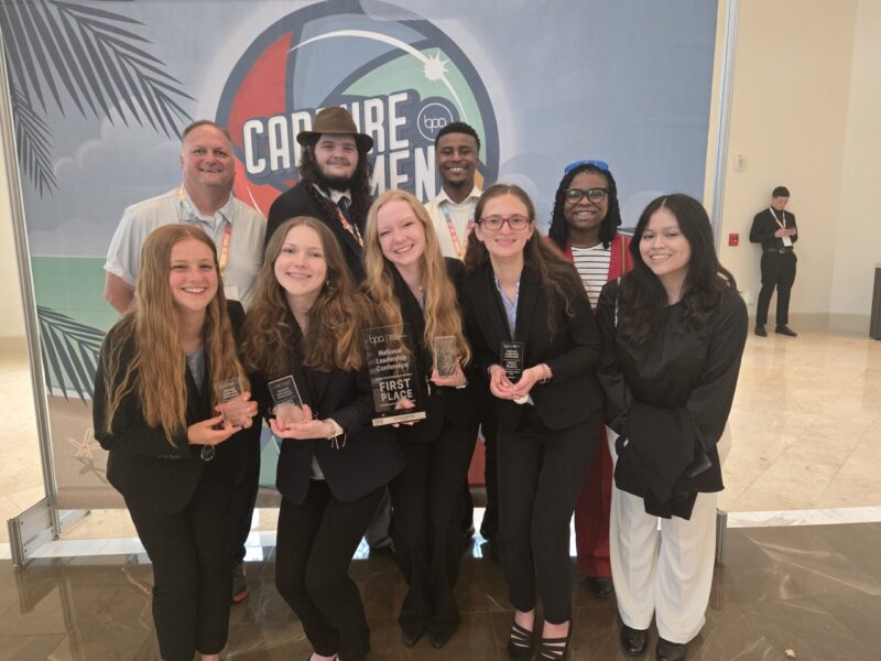 A group of eight smiling young adults, some holding trophies, stand together in front of a 