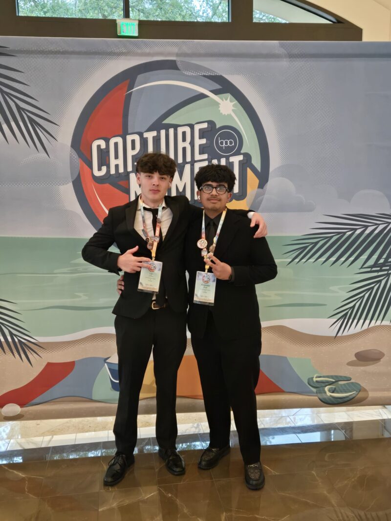 Two young men wearing black suits and conference badges stand arm-in-arm in front of a colorful 
