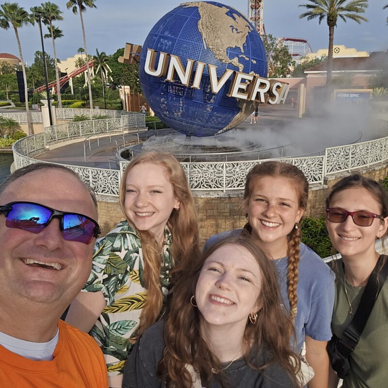 A man and four young women, including a BPA National Champion, smile for a selfie in front of the large Universal Studios globe entrance, surrounded by palm trees and mist on a sunny day.
