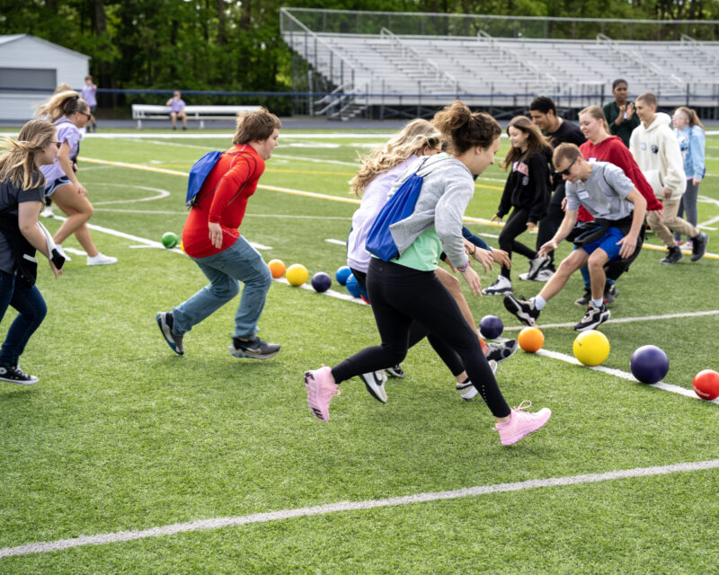 A group of teenagers at Edgewood run toward colorful balls on a sports field, seemingly starting a game, with bleachers and trees in the background.