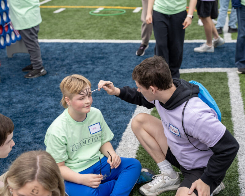 A person kneels on artificial turf while painting a child's face at an Edgewood outdoor event. The child, wearing a light green shirt, smiles as others enjoy games in the background—capturing the lively spirit of the Ross and DECA community.