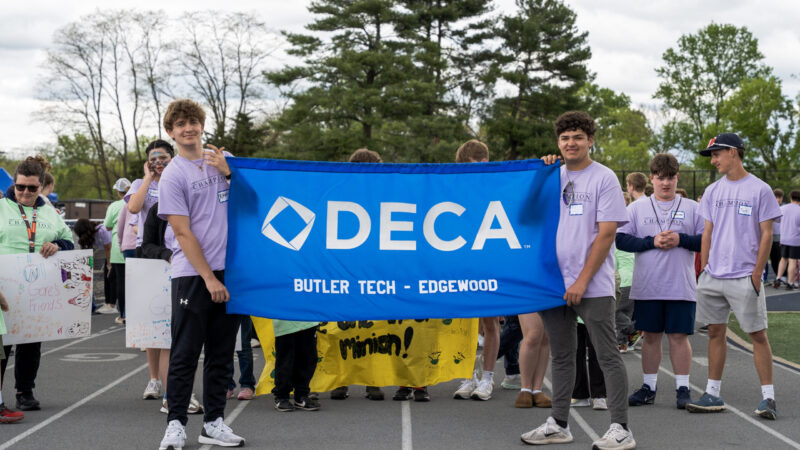 A group of students in light purple shirts hold a blue banner that reads