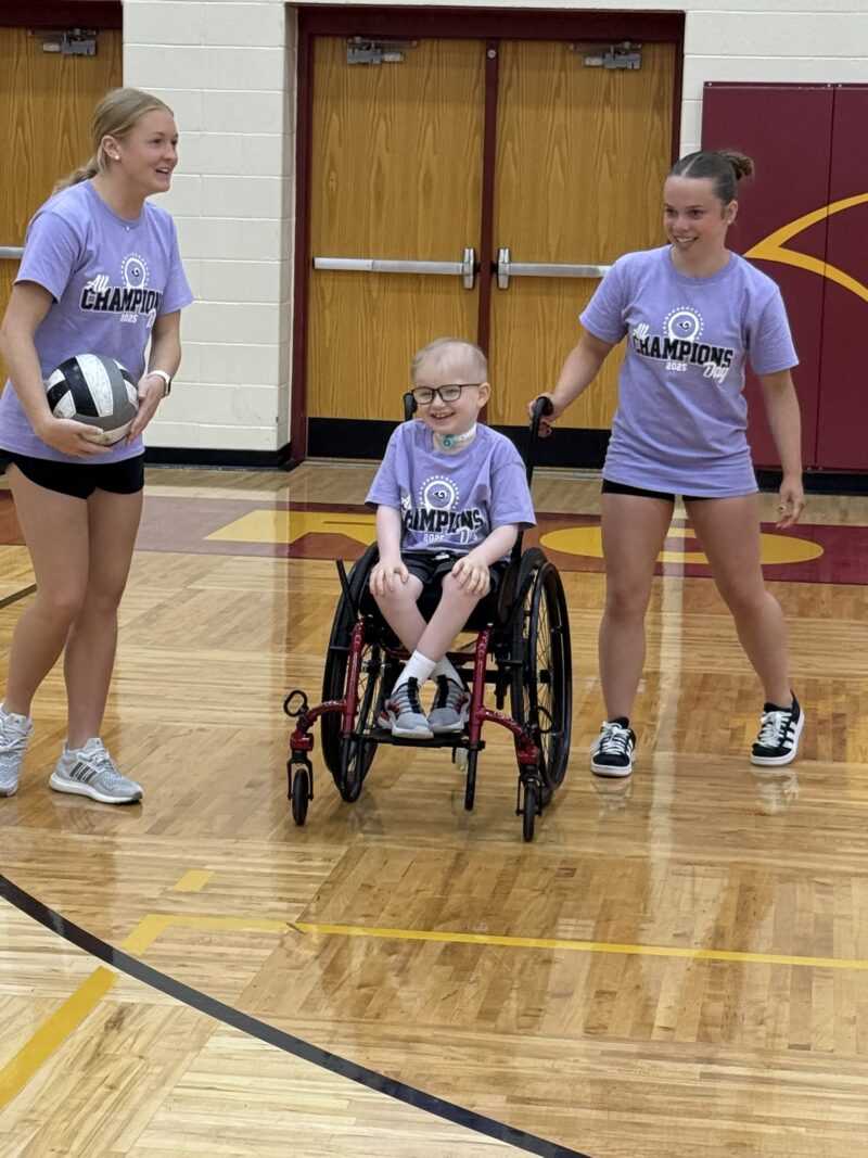 Two young women from Edgewood stand in a gym with a child in a wheelchair between them. All are smiling and wearing matching purple