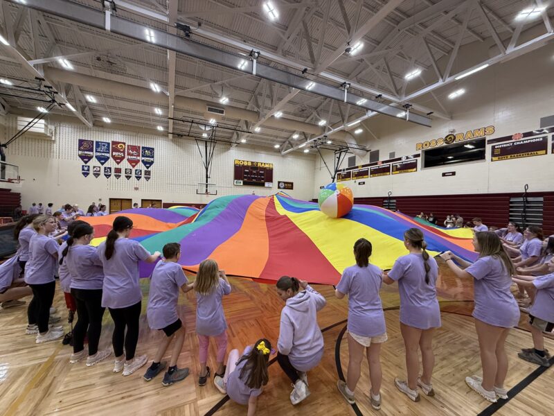 Children in matching purple shirts from Edgewood hold the edges of a large, colorful parachute in a gymnasium, bouncing a giant beach ball on top as part of a group activity.