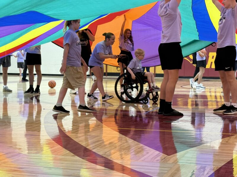 Children in gym clothes lift a colorful parachute in the Edgewood gymnasium, with one child in a wheelchair smiling as they join the group. A basketball sits on the floor in the background.