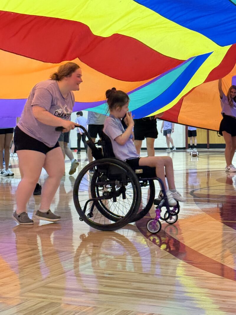 In the Edgewood gymnasium, a woman pushes a girl in a wheelchair under a large, colorful parachute. Both are smiling and participating in an inclusive group activity with others enjoying the fun in the background.