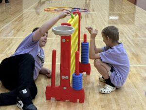 Two boys in purple shirts sit on the gym floor at Edgewood, playing with a colorful vertical toy. Focused on the activity, these Champions of All Abilities enjoy their time together while hoops rest in the background.