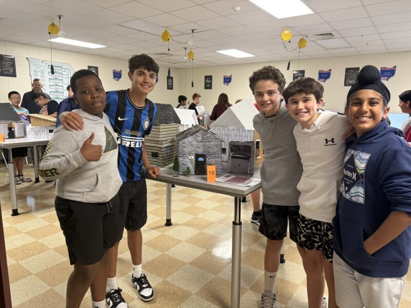 Five smiling boys stand together in a Junior High classroom behind their Tiny House Challenge model project, proudly displaying their work as young designers among other students and creative displays in the background.