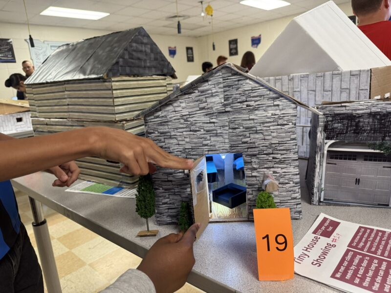 A person points at a detailed Tiny House model with a stone exterior, lit interior, and small bushes by the door. The model is part of the Tiny House Challenge for Junior High Designers, displayed with card 19 and informational sheets.