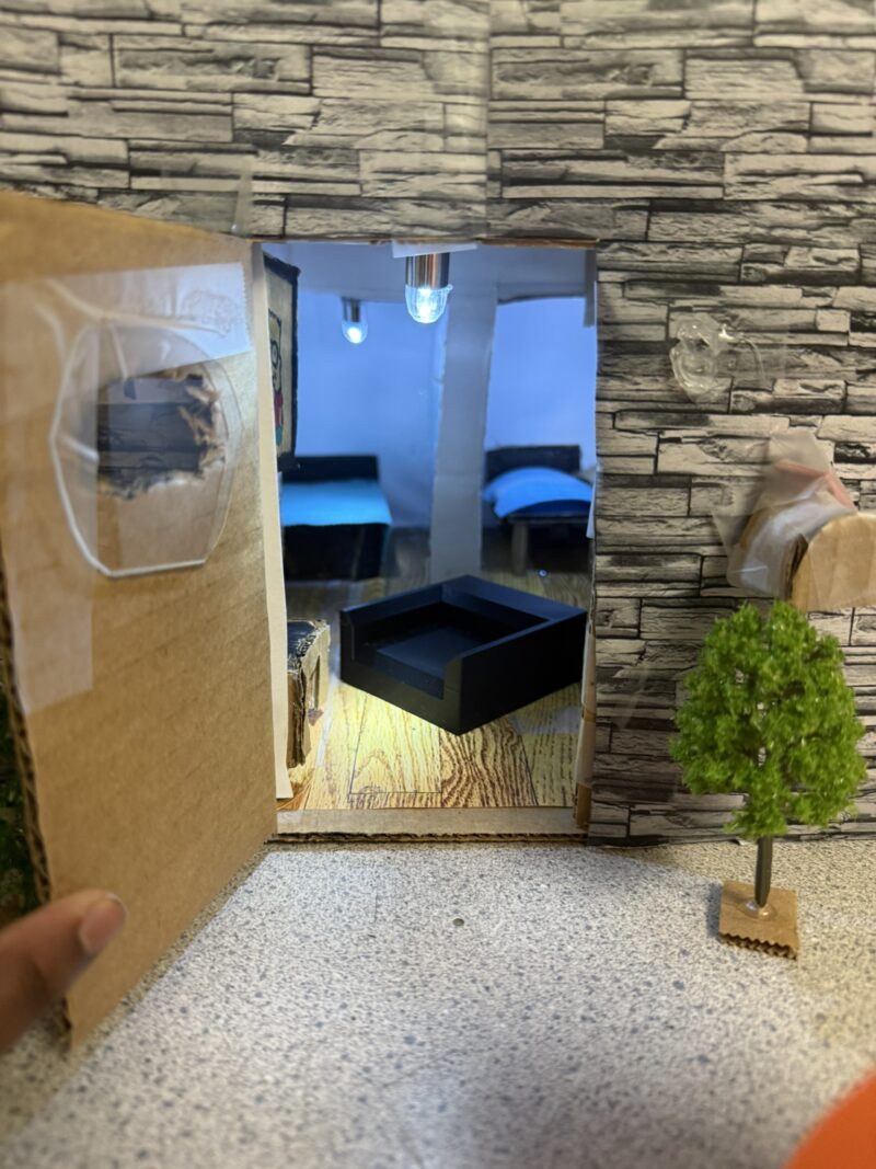 A miniature Tiny House with a stone-patterned exterior, open cardboard door, small tree outside, and two beds with blue blankets inside under a ceiling light—perfect for a Junior High Designers’ Tiny House Challenge.
