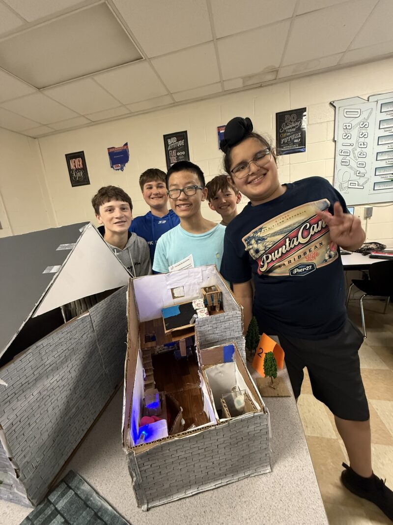 Five smiling children stand behind a detailed Tiny House model in a classroom, posing for the photo; one child in front flashes a peace sign. Classroom posters and desks are visible, showcasing the Junior High Designers’ latest creation.