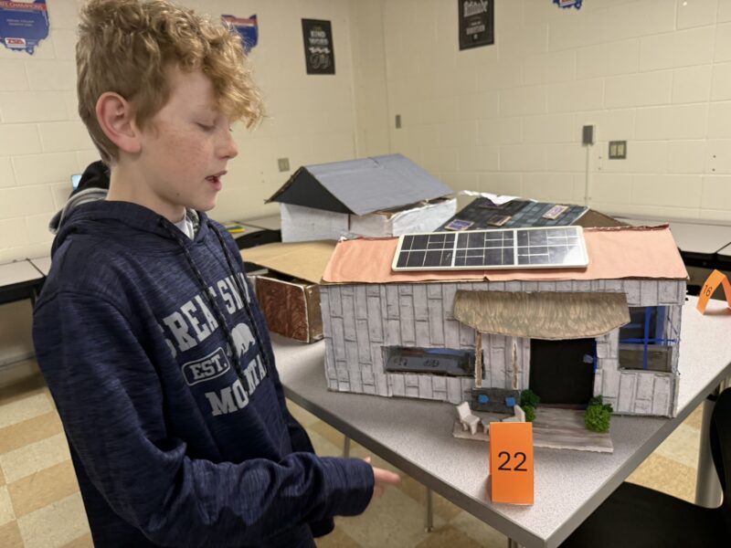 A boy with curly hair stands beside his Tiny House model with a solar panel on its roof, displayed on a classroom table and marked with the number 22—one of many creative entries in the Junior High Designers’ Tiny House Challenge.