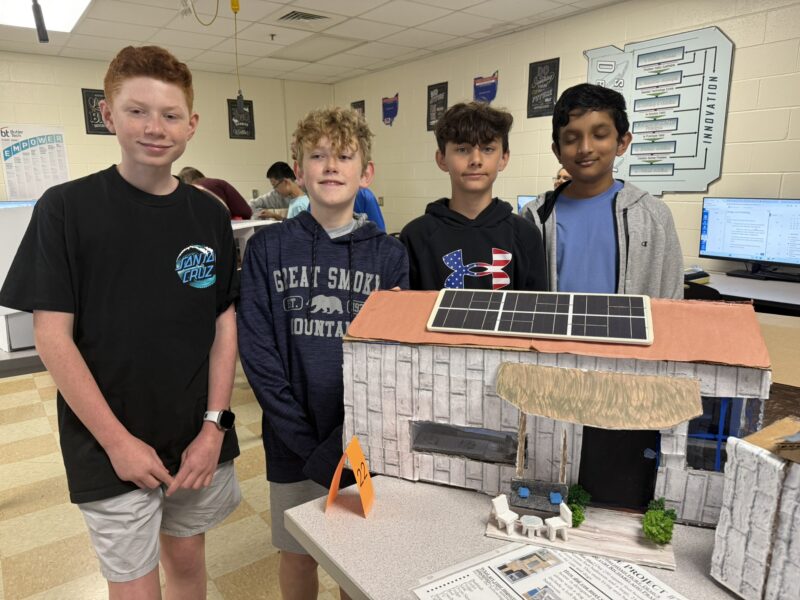 Four junior high designers stand together in a classroom, smiling beside their detailed model for the Tiny House Challenge, featuring solar panels. Posters and computers fill the background, highlighting their creative efforts.