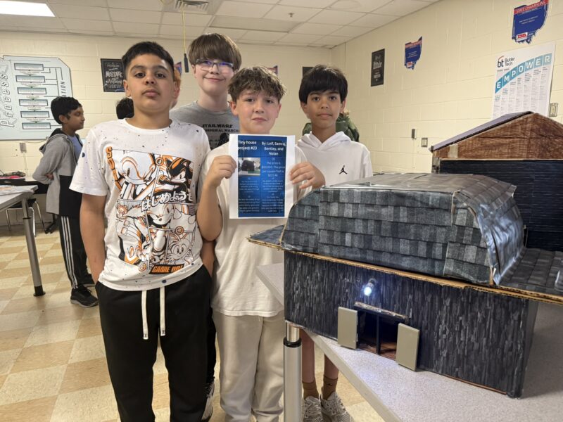Four boys stand in a classroom next to a detailed model for the Tiny House Challenge. One boy holds a project sheet with text and an image, while other students and classroom posters provide design inspiration in the background.