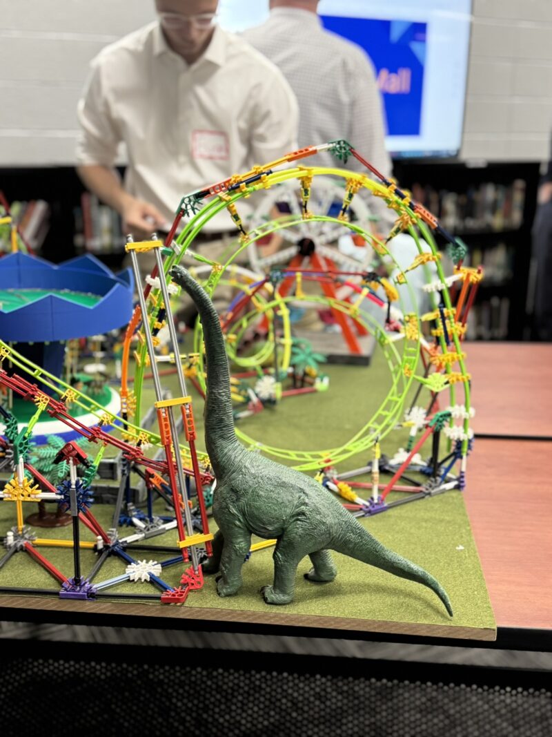 A green toy dinosaur stands in front of a colorful K'NEX roller coaster model on a table at the Butler Tech Engineering Capstone Showcase, with two people blurred in the background.