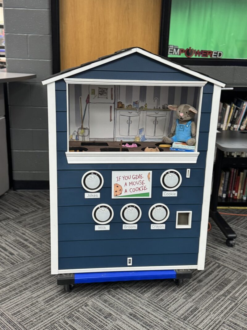 A blue and white house-shaped display at the Lakota East Engineering Capstone Showcase features a mouse doll in a kitchen scene with a sign reading